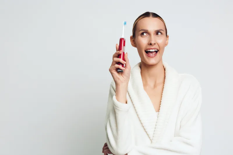Smiling woman in white bathrobe holding red electric toothbrush with toothpaste on bristles. Clean background, concept of dental hygiene, oral care, and morning routine. Bright lighting.