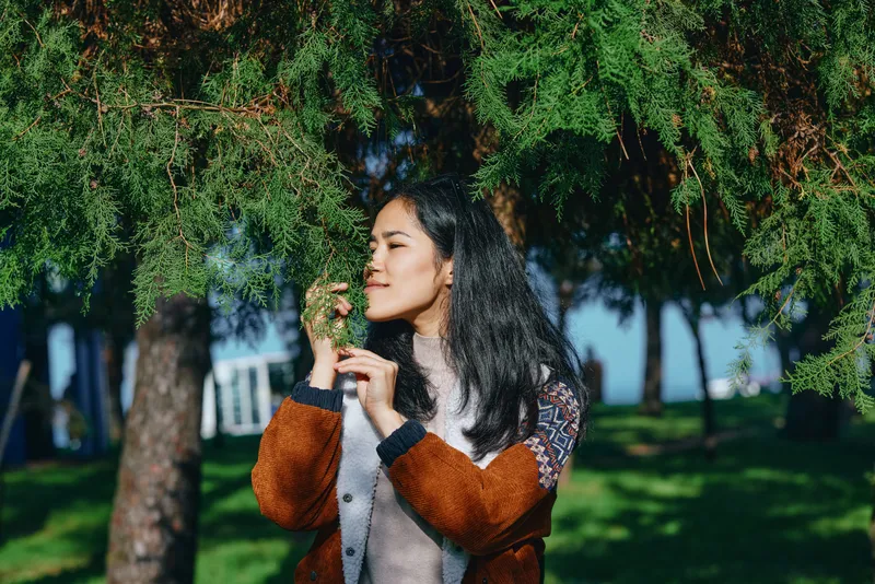 Woman enjoying nature outdoors in a park, smelling a flower beneath leafy branches, wearing a cozy cardigan, sunlight creates a serene lifestyle moment that conveys calm, wellness, and connection