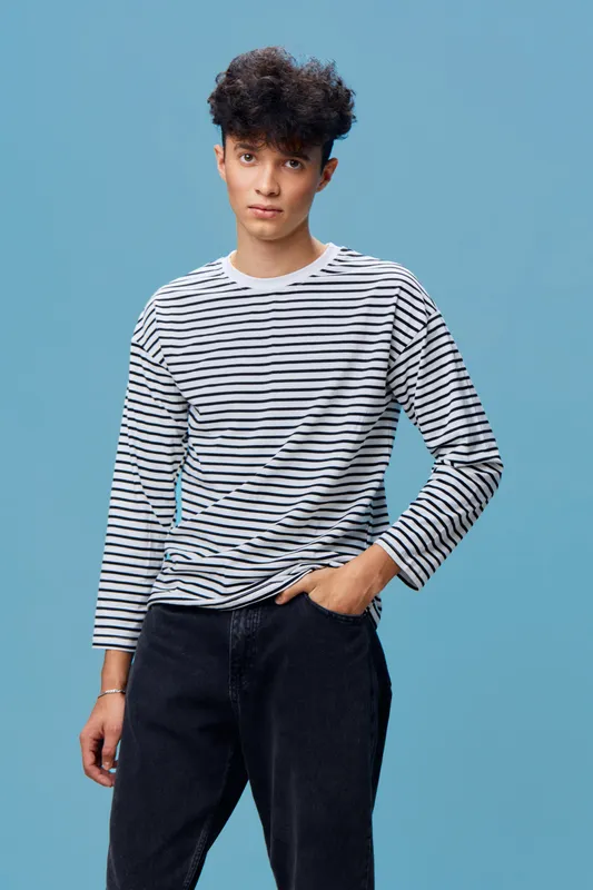 Young man in a striped long-sleeve shirt standing relaxed against a blue background, casual style, dark jeans, curly hair, confident and calm expression.