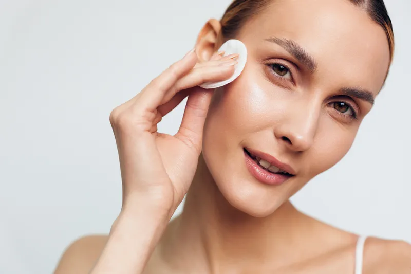 Young woman with clear skin removing makeup using cotton pad, gentle skincare routine, natural beauty, close-up portrait, soft lighting, clean background, fresh and healthy appearance