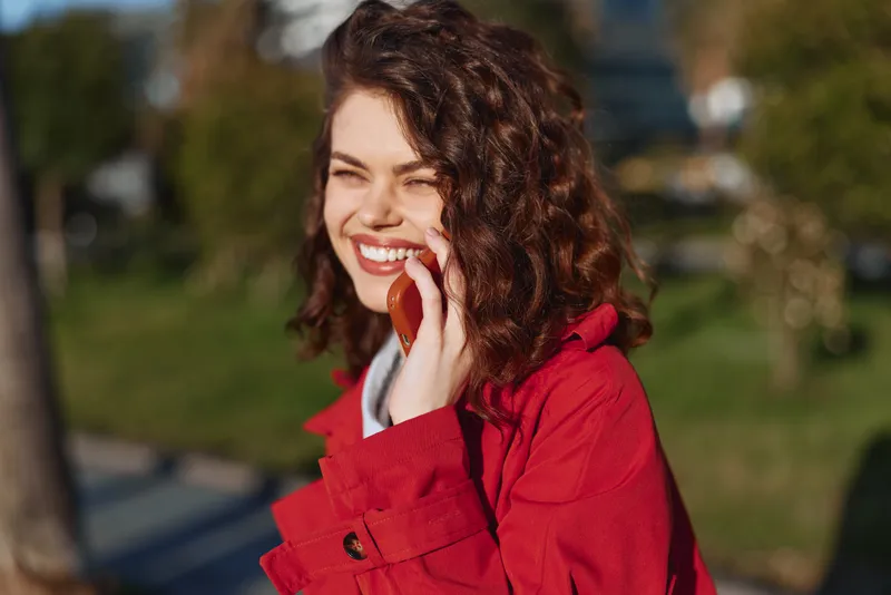 A smiling woman in a red coat chats on a phone in a sunny park, standing among trees with soft afternoon light, embodying casual connection and contemporary confidence.