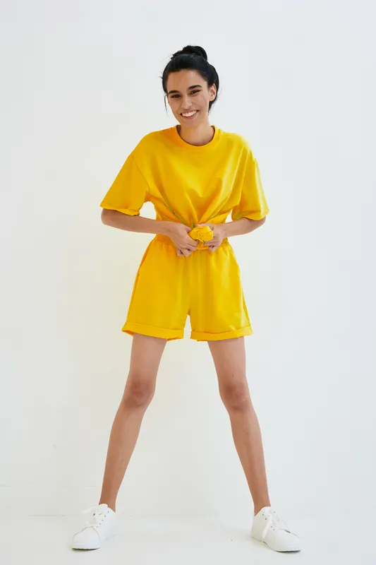 Young woman wearing bright yellow casual shorts and t-shirt, smiling confidently, posing against a clean white background, styled for summer comfort and active lifestyle.