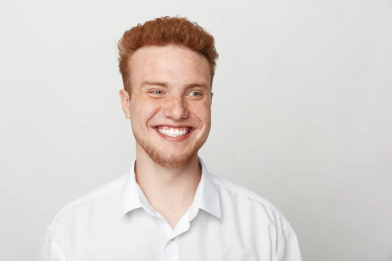 Smiling young man with red hair and beard wearing white shirt looks away on light background. Portrait of happy male model with natural expression and casual style in studio lighting.