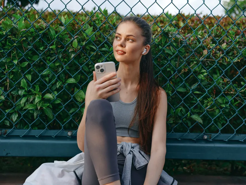 Расслабленная молодая женщина наслаждается музыкой на смартфоне, сидя на скамейке у зелёной кустарниковой изгороди, одетая в спортивную одежду