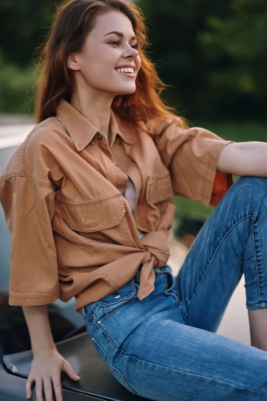 Young woman in casual outfit exuding joy outdoors, wearing a brown oversized shirt and blue jeans, with a lush green background.