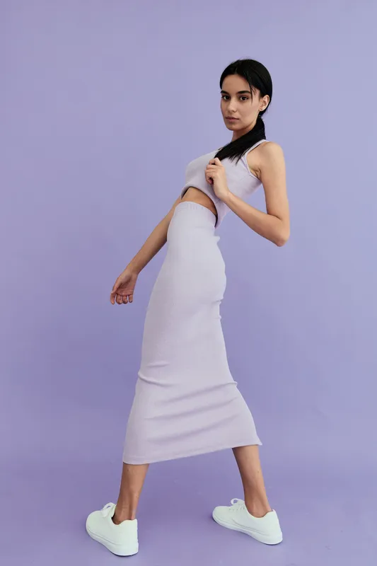 Young woman in a lilac knitted dress poses confidently against a solid pastel purple background, wearing white sneakers and minimalist makeup for a modern casual look.
