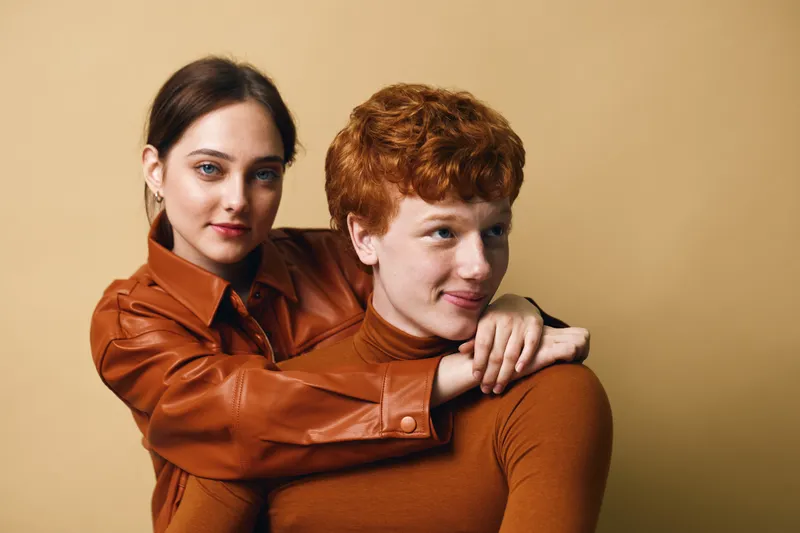 Young woman with blue eyes and brown hair gently hugs redhead man wearing brown turtleneck, studio portrait with warm beige background and cozy fashion style.
