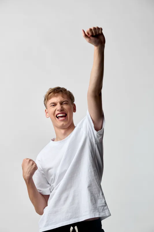 Energetic victory pose, man celebrating with raised fist and broad smile, casual white tee, dynamic gesture conveying triumph, motivation, and festive success