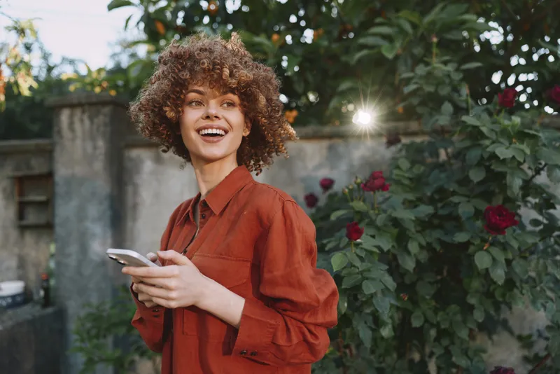 Cheerful woman outdoors, smiling with curly hair, holding phone in a sunlit garden, casual fashion with natural colors, blooming flowers, candid mood, relaxed and hopeful