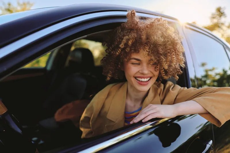 A young woman with curly hair leans out of a car window, smiling in warm retro tones for a candid lifestyle moment.