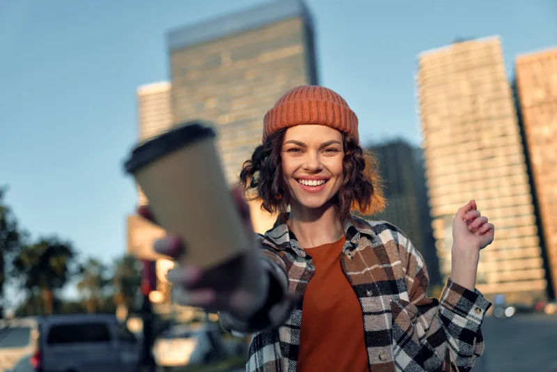 Woman offering coffee in city outdoors, beanie and flannel, smile and casual vibe, candidlifestyle authenticity with goldenhourglow, mindful living and emotionalstorytelling atmosphere.