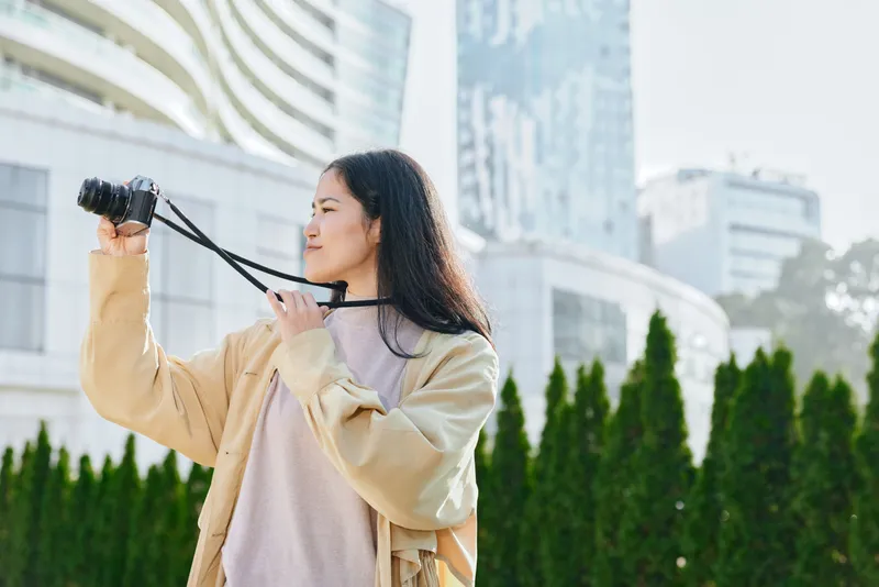 Woman capturing city life with a camera outdoors, urban backdrop and modern architecture, showcasing candid travel moments and creative storytelling in daylight