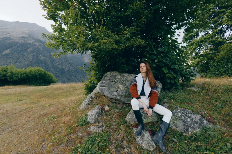 Woman nature mountains tree rock meadow hiking boots portrait of a young redhead sitting on a boulder in countryside, wearing cableknit sweater and rubber boots for a serene outdoor escape.