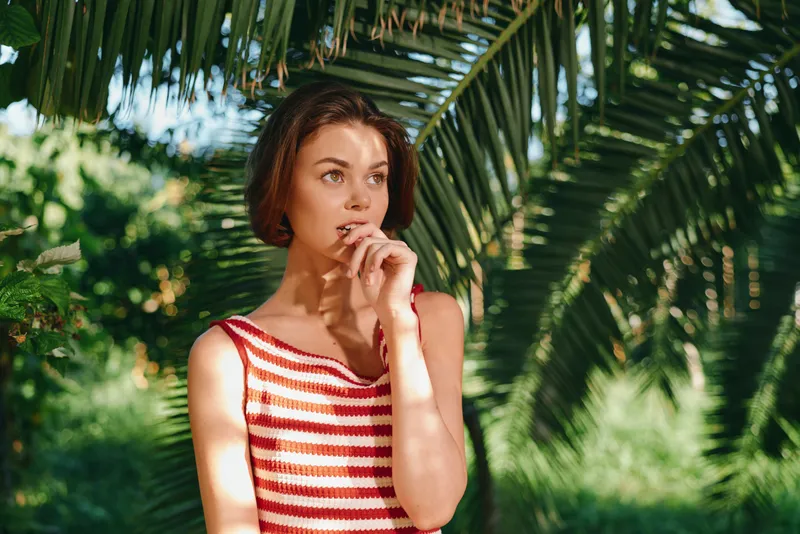 A thoughtful woman stands among lush greenery, wearing a striped sleeveless top. She gazes away, lost in thought, conveying calm, curiosity, and natural charm in bright outdoor light.