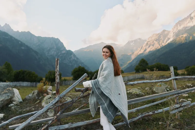 Young woman in a gray poncho smiles over her shoulder as she stands beside a wooden fence in a sunlit mountain valley