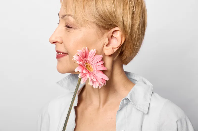 Woman portrait closeup, blonde woman holding pink flower near ear, calm serene expression, soft light, white background, casual shirt, fresh spring beauty