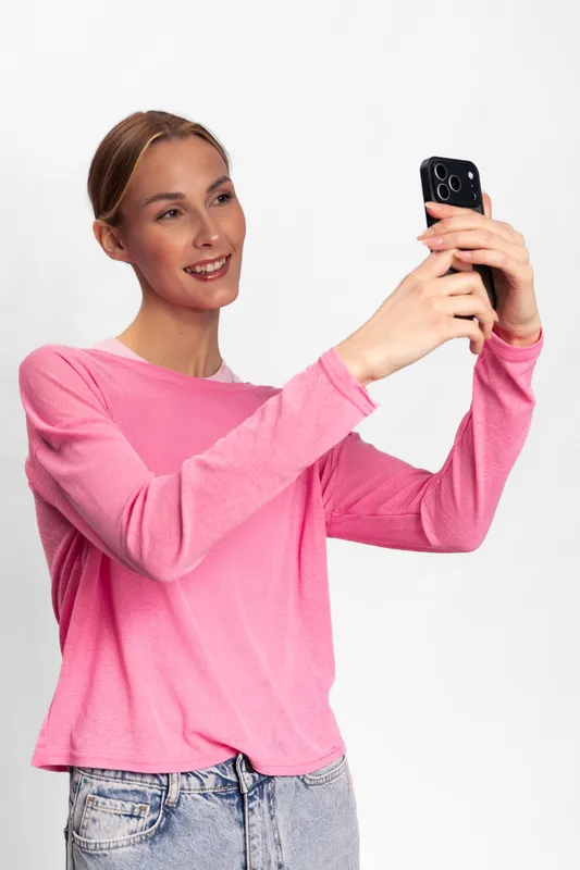 Young woman in pink sweater smiles while taking a selfie with smartphone. Casual style, natural light, isolated on white background. Concept for social media, technology, and youth culture.