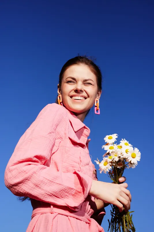 Joyful young woman in a pink dress holding daisies against a vibrant blue sky, radiating happiness and positivity in springtime