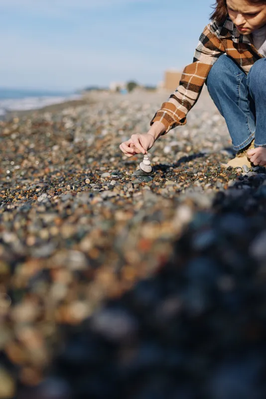 Casual person kneeling on a pebble beach, wearing a plaid shirt and jeans, collecting small stones near the shoreline with the sea in the background and coast