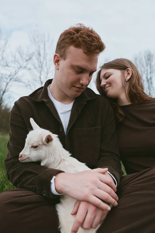 Couple cradles a white goat kid outdoors, showcasing affectionate companionship in a casual lifestyle portrait. Soft natural light and rustic backdrop convey warmth, care, and rural charm.
