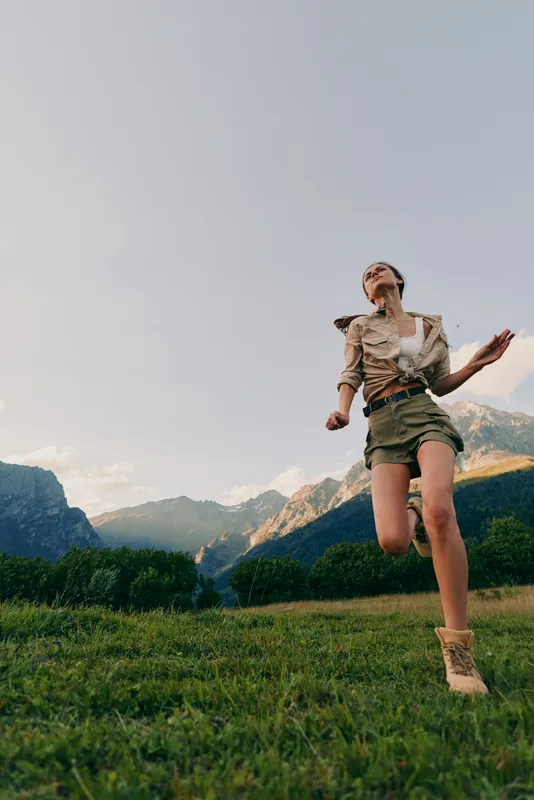 Young woman strolls across a sunlit alpine meadow with distant mountains, feeling freedom and joy as she moves in hiking boots under a clear sky.