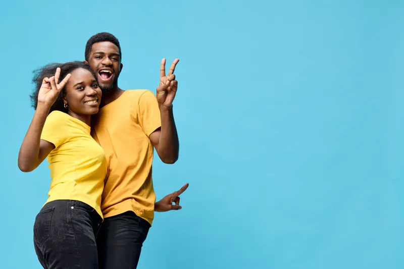 Young African American couple in matching yellow shirts, smiling joyfully against a bright blue background, expressing love and happiness with peace signs