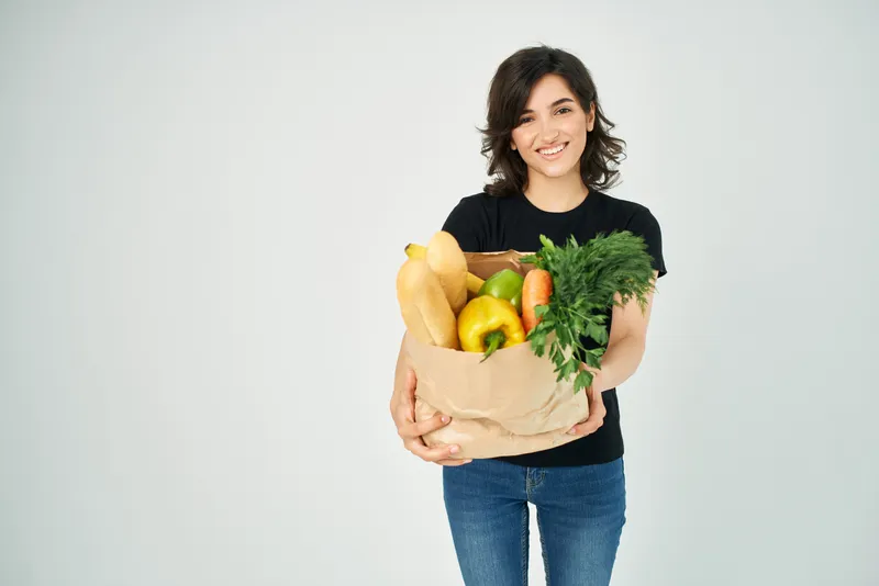 Весёлая женщина с пакетом продуктов, овощами, здоровой пищей на светлом фоне