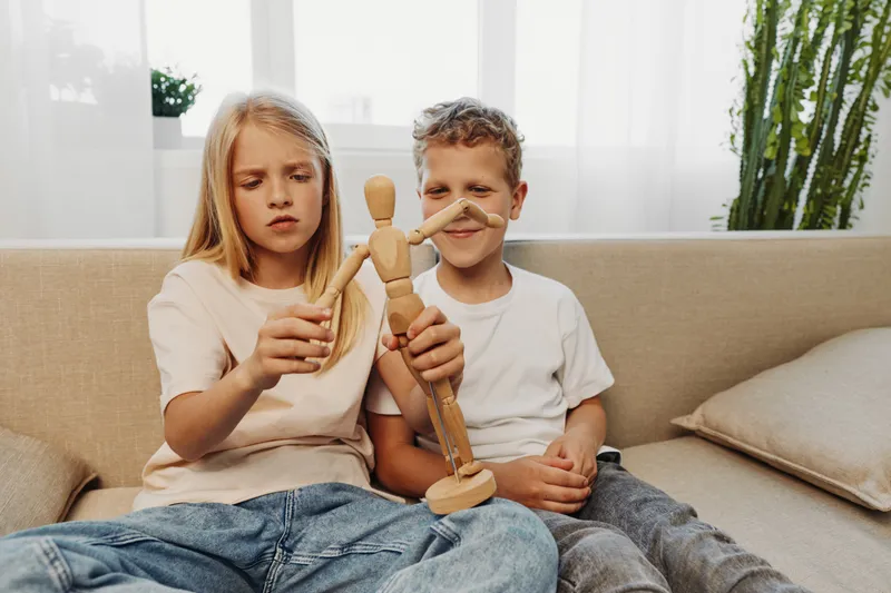 Two children sitting on a beige couch holding a wooden mannequin toy, showing curiosity and concentration in a bright room with natural light and indoor plants.