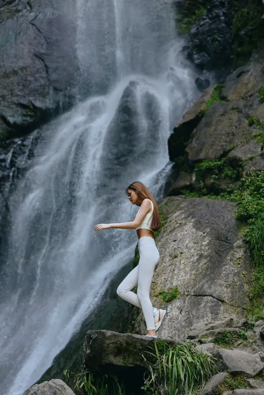 Woman in white activewear walking near a cascading waterfall, surrounded by lush green rocks and nature, showcasing outdoor adventure and fitness lifestyle.
