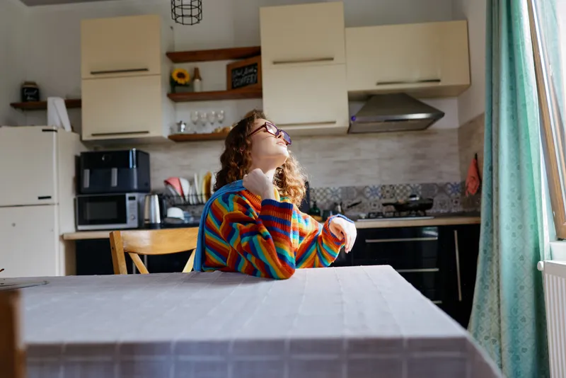 A woman in a colorful striped sweater sits at a kitchen table, gazing upward with a thoughtful smile. Bright morning light fills a modern home kitchen scene.