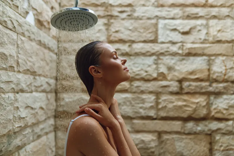 Woman enjoying warm shower under rainfall showerhead in rustic stone bathroom with natural light, conveying relaxation and peacefulness in a serene spa-like environment.