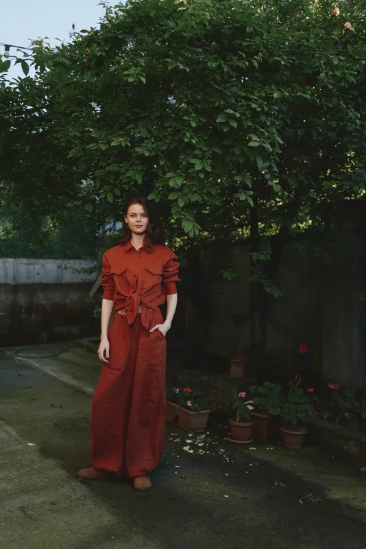 Woman outdoors, standing in a garden wearing a rust red outfit, relaxed pose, soft sunlight, natural colors, lush greenery background, lifestyle moment