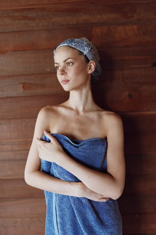 young woman wrapped in blue towel with hair net standing against wooden wall, natural light, serene expression, wellness, spa atmosphere, calm mood