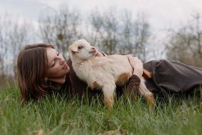 Loving woman lying on grass with a baby goat, outdoor scene showing gentle bond and lifestyle. Soft natural light creates a warm, tender mood of care and happiness.