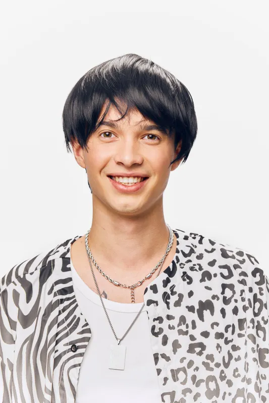 smiling young man with black hair wearing patterned shirt and layered necklaces on white background, studio portrait with cheerful expression