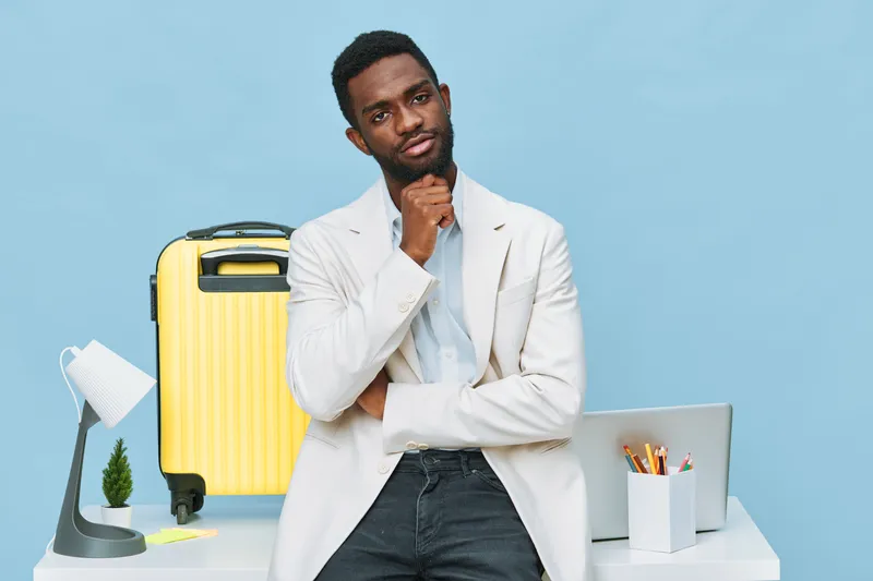 Thoughtful man in smart casual attire stands at modern workspace with laptop and yellow suitcase, contemplating decision. Bright minimalist background emphasizes professional and travel readiness. Peo