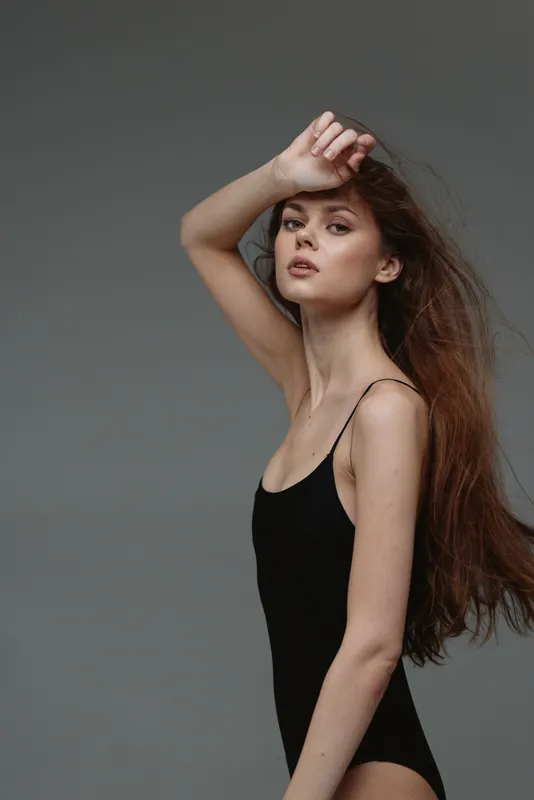 Elegant woman with long hair wearing a black bodysuit posing against a gray background, showcasing natural beauty and confidence.