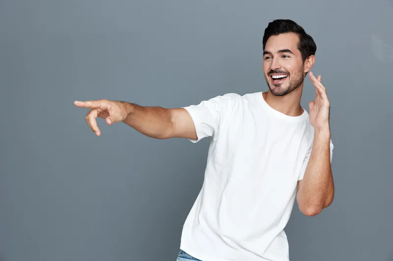 Smiling man wearing a casual white t-shirt looking joyfully at the camera while gesturing with his hand against a solid grey background. Lifestyle concept.