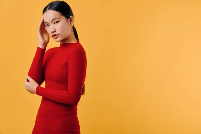 Young woman in red sweater with long sleeves poses thoughtfully against orange background. Model with long dark hair looks down with hand near head in studio shot.