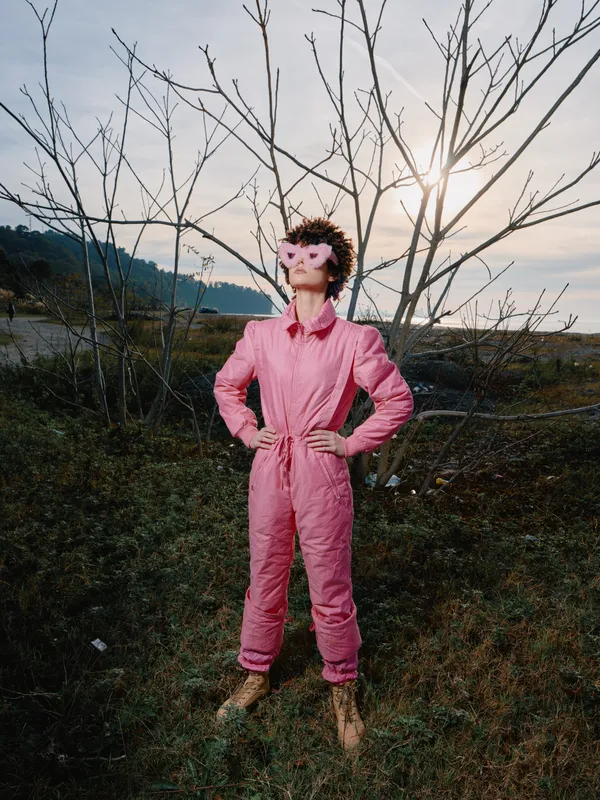 Confident person in a pink jumpsuit outdoors hands on hips sun glow behind leafless trees rugged landscape fashion portrait with bold color and strong stance