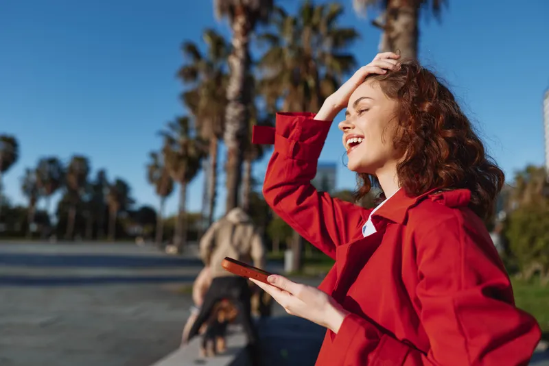 Young woman with curly hair enjoys a sunny day outdoors while smiling and holding a smartphone near palm trees.