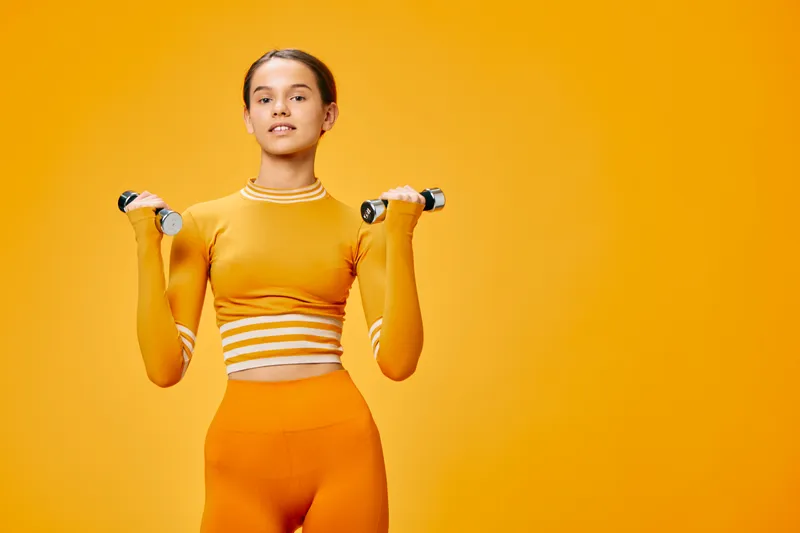 Woman fitness enthusiast with dumbbells in an orange studio, wearing a cropped top and leggings, demonstrating strength, balance and focus during a dynamic workout session.