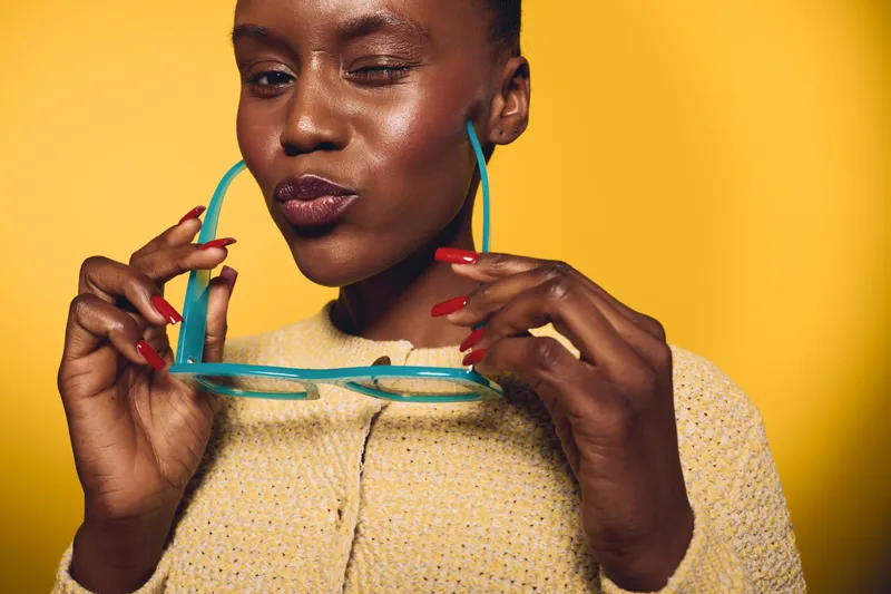 Confident dark skinned African American woman winks holding turquoise sunglasses with red nails in front of yellow background. Concept of fashion, beauty, summer and playful mood.