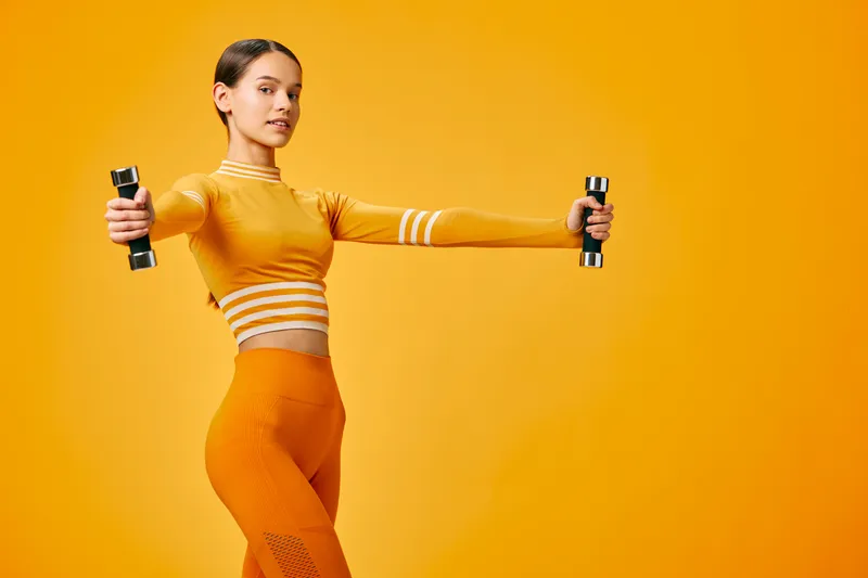Fitness-minded woman performs a dynamic workout with dumbbells in a bright orange outfit, showcasing athletic form and balance against a bold orange backdrop.