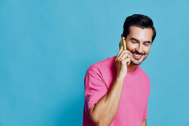 Smiling man in casual pink t-shirt holds smartphone, looking cheerful while talking on the phone against a vibrant blue background. Modern lifestyle concept.