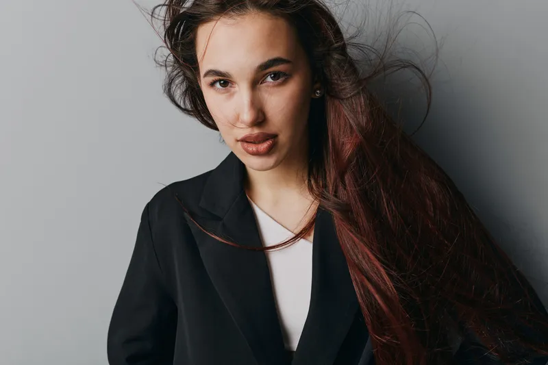 Young woman with long hair wearing black blazer and white shirt posing against gray background with confident expression and natural makeup in studio portrait session.