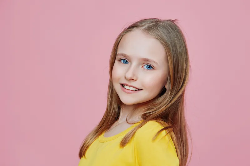Cheerful young girl with long blonde hair wearing a yellow shirt, smiling against a soft pink background, embodies joy, innocence, and youthfulness