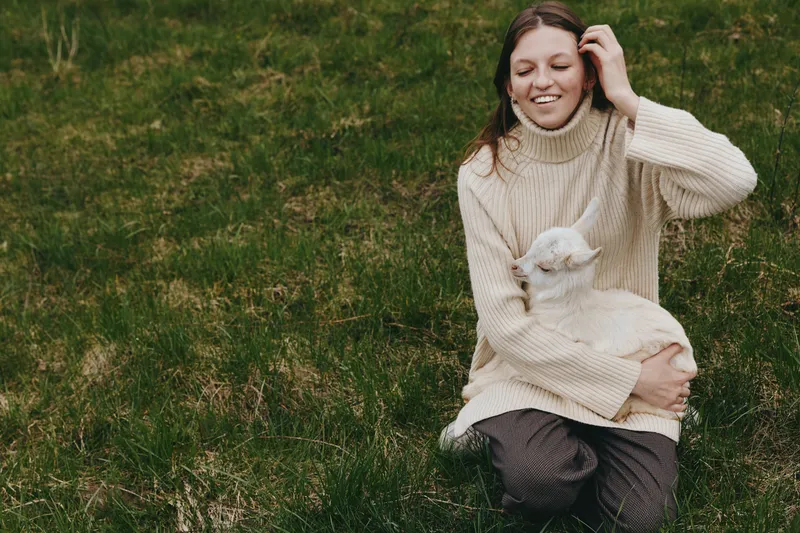 Cheerful woman outdoors, embracing a gentle goat kid in her arms, sunlit grass around, cozy sweater, relaxed posture, natural colors, peaceful countryside moment, warm connection