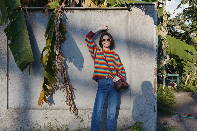 Woman in striped sweater and sunglasses smiling while leaning against a concrete wall in tropical outdoor setting, casual outfit with blue jeans and shoulder bag for travel lifestyle and fashion.