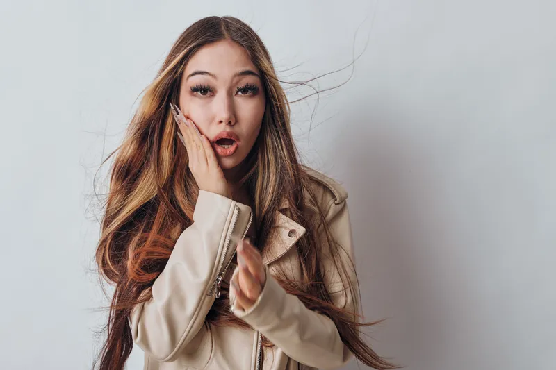 Young woman with long hair in beige jacket showing surprised expression touching face with open mouth and looking at camera on neutral background.